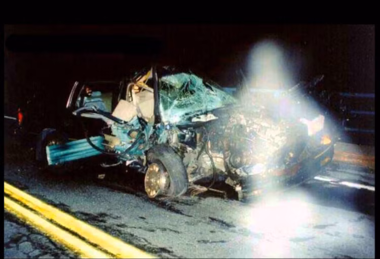 Photograph of a spectral figure in front of a ruined car.